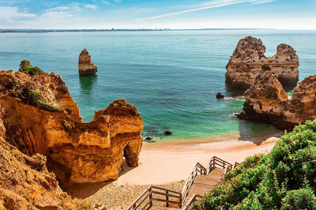 The image captures a scenic beach in Algarve, Portugal. The beach features golden sandy shores with a clear pathway leading down to the water. The ocean is a vibrant turquoise, and several large rock formations rise from the water near the shore. Lush greenery borders the top edge of the beach, and in the distance, a calm horizon meets a clear blue sky
