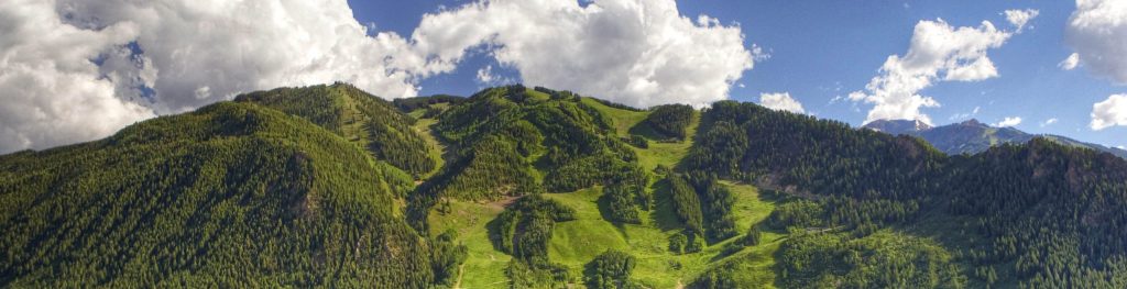 The image shows a breathtaking view of lush green mountains, in Aspen Mountain range, with a town nestled at the base. The mountains are covered with dense forests, and the slopes are clearly visible with patches of open greenery. The sky is a deep blue with large, fluffy white clouds floating above the peaks. The sunlight casts a warm glow over the landscape, highlighting the natural beauty of the scene. The town below appears peaceful, surrounded by trees and greenery, blending harmoniously with the natural environment.