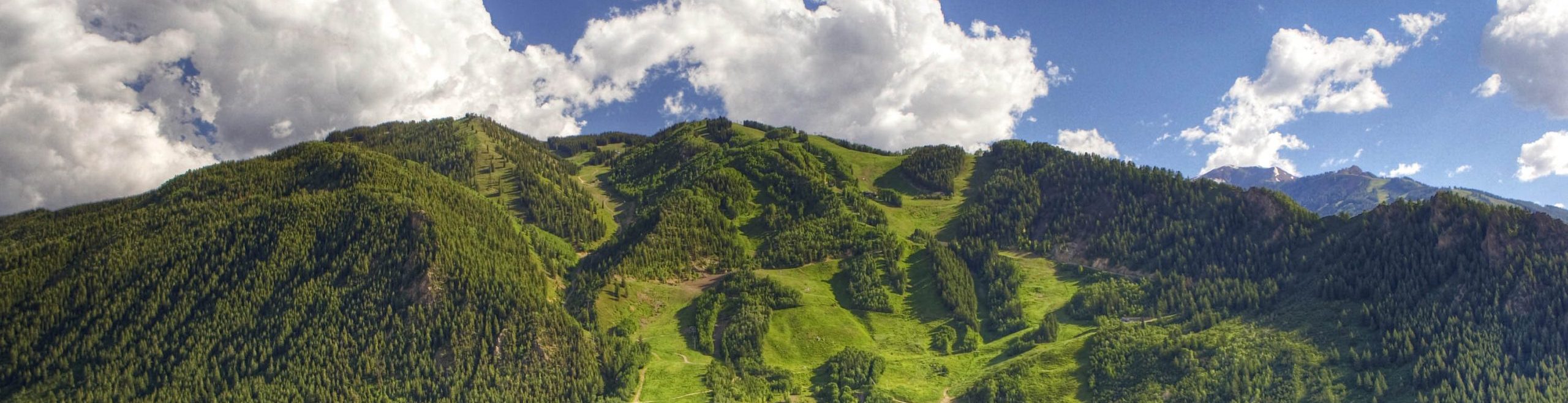 The image shows a breathtaking view of lush green mountains, in Aspen Mountain range, with a town nestled at the base. The mountains are covered with dense forests, and the slopes are clearly visible with patches of open greenery. The sky is a deep blue with large, fluffy white clouds floating above the peaks. The sunlight casts a warm glow over the landscape, highlighting the natural beauty of the scene. The town below appears peaceful, surrounded by trees and greenery, blending harmoniously with the natural environment.