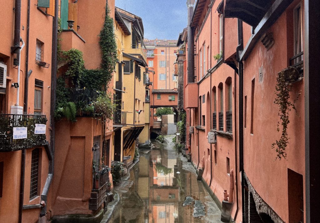 This photo captures the charming view of the hidden canal from the Finestrella di Via Piella in Bologna, often referred to as the city's "Little Venice." The canal, lined with brightly colored terracotta buildings in shades of orange and red, reflects the vibrant architecture and the serene, narrow waterway running between them. The tranquil scene showcases the contrast between the peaceful water and the urban structures above, with shuttered windows, small balconies, and green plants clinging to the buildings. It's a rare and picturesque glimpse into Bologna's historical canal system, a remnant of the city's rich past.