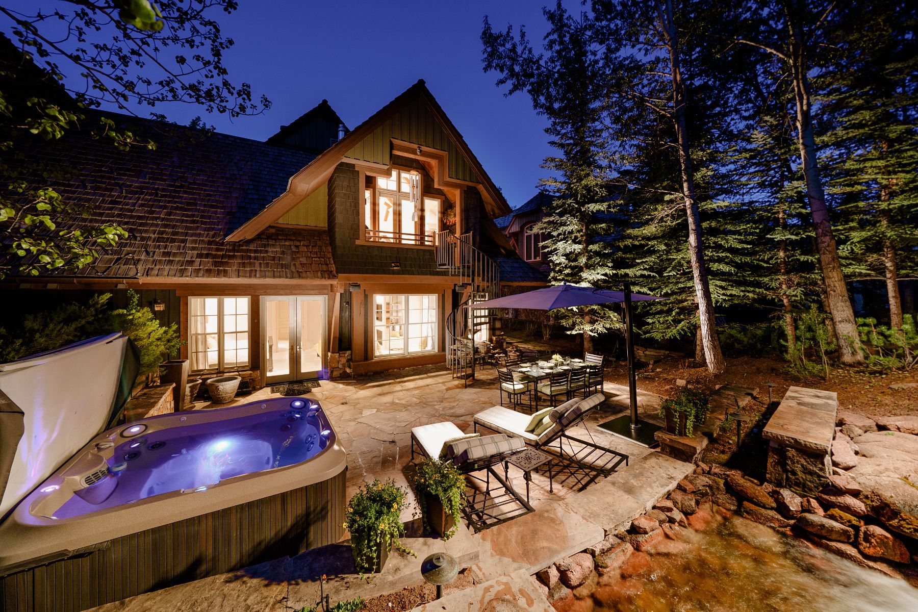 An image of a luxurious chalet in Aspen during twilight. The two-story structure features a steep gabled roof with exposed wooden beams and large windows emitting warm light. In the foreground, there is an inviting outdoor hot tub on a stone patio, alongside outdoor furniture under an open-air shelter with a fabric canopy. Surrounding the chalet are tall pine trees that add to the serene mountain retreat ambiance.