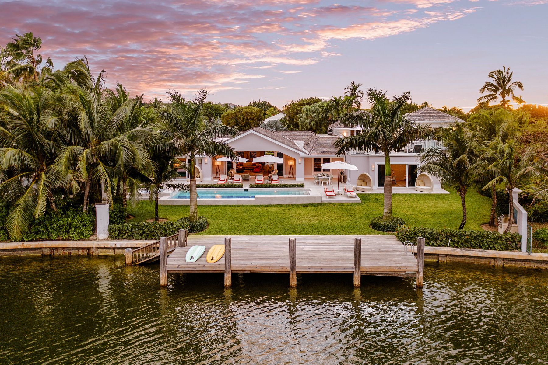 Image of one of our homes. A luxurious waterfront villa in the Bahamas during twilight. The image showcases a large, elegant house with a terracotta roof and white walls, surrounded by lush green palm trees. In front of the villa is a spacious patio area with an outdoor pool, lounge chairs, and an open-air seating area. A wooden dock extends into the calm water with two paddleboards resting on it, highlighting the recreational opportunities available. The serene atmosphere is enhanced by the soft glow of lights from within the house and around the pool area, suggesting a peaceful evening setting.