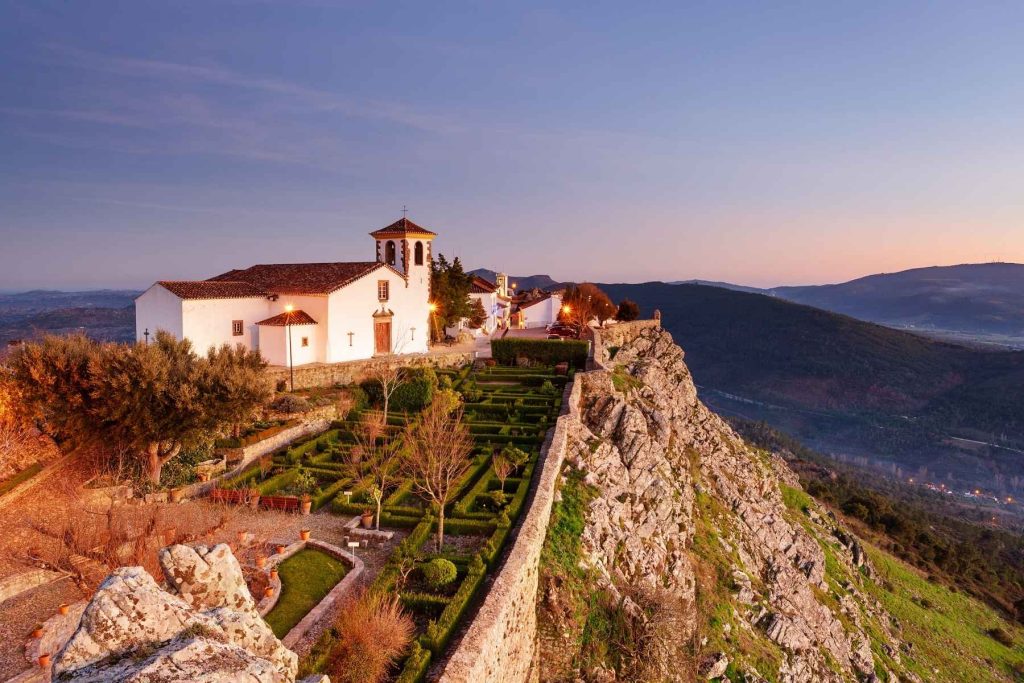 A panoramic view of Marvão, a picturesque village perched on a granite crag at the top of a high hill. The image features a prominent white building with terracotta roofing and a bell tower, illuminated by warm lights against the twilight sky. In the foreground, there are terraced gardens and rugged rock formations, while in the background, layers of rolling hills fade into the horizon under a soft purple sky. This is one recommendation for the Alentejo Itinerary.