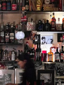 The photo captures a dimly lit bar or café with shelves filled with a variety of bottles and memorabilia. The shelves are stocked with different types of liquor bottles, including brands like Havana Club, Carpano, and Jägermeister. Mixed among the bottles are personal items such as photographs, books, and small decorative pieces, adding a nostalgic and eclectic atmosphere to the scene. Black-and-white photographs and quirky objects give the space a vintage, artsy vibe. In the foreground, there is a blurred figure, likely a person in motion, creating a dynamic contrast to the stillness of the bar's backdrop. The overall mood is cozy and intimate, likely representing a popular local spot