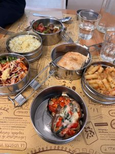 The photo displays a meal served in small metal containers, arranged in a bento-style manner. The dishes include a baked eggplant topped with melted cheese, spinach, and diced tomatoes; a serving of pasta in a light tomato sauce; a portion of seasoned rice; a fresh mixed vegetable salad with grated carrots, cabbage, and greens; and what appears to be sautéed vegetables or a stew. Additionally, a piece of flatbread or focaccia is served alongside. The setting is casual, with glasses of water and a placemat featuring food-related texts and illustrations, evoking a simple, healthy, and rustic dining experience, likely from a vegetarian or health-conscious restaurant.