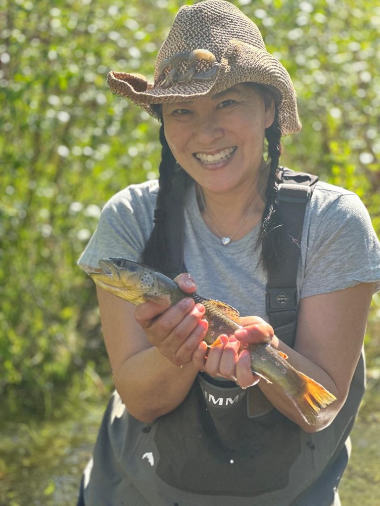 In the photo, Cathy stands proudly holding a freshly caught fish, cradling it with both hands. She beams with excitement, showcasing her catch to the camera. The fish glistens in the sunlight, its scales reflecting shades of silver and green. The woman is dressed in outdoor or fishing gear, wearing a hat. In the background, you can see a natural setting, a river, lake, surrounded by lush trees or mountains, completing the scene of a successful fishing trip. The atmosphere is joyful and adventurous