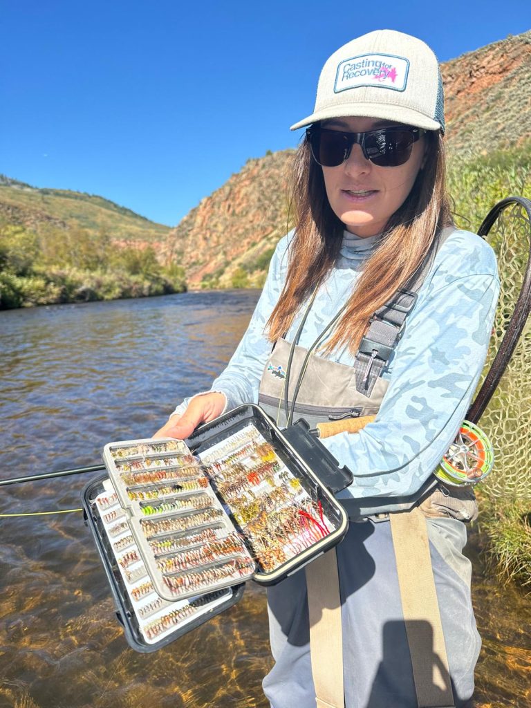 In the photo, the fishing guide stands near the edge of a tranquil lake in Aspen, holding fishing gear. Dressed in outdoor apparel, including waders, the guide is prepared for the day’s fishing tour. The backdrop of the lake is stunning, with the calm water reflecting the surrounding mountains and autumn-colored trees. The guide’s posture suggests they are ready to lead the group on the tour, with a sense of expertise and familiarity with the beautiful natural surroundings. The atmosphere feels peaceful and inviting, with the scenic Aspen landscape enhancing the outdoor adventure.