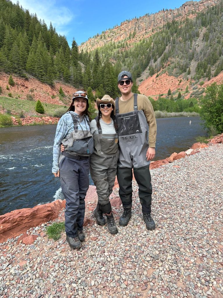 The photo shows Cathy and Brandon standing in a river alongside their fishing guide. All three are dressed in fishing gear, including waders and hats. The guide, standing onde their left side, seems be joyful. The river surrounds them, reflecting the sunlight, and the natural environment is peaceful, with trees or mountains visible in the background. The scene captures a shared moment of learning and enjoyment during a guided fishing trip.