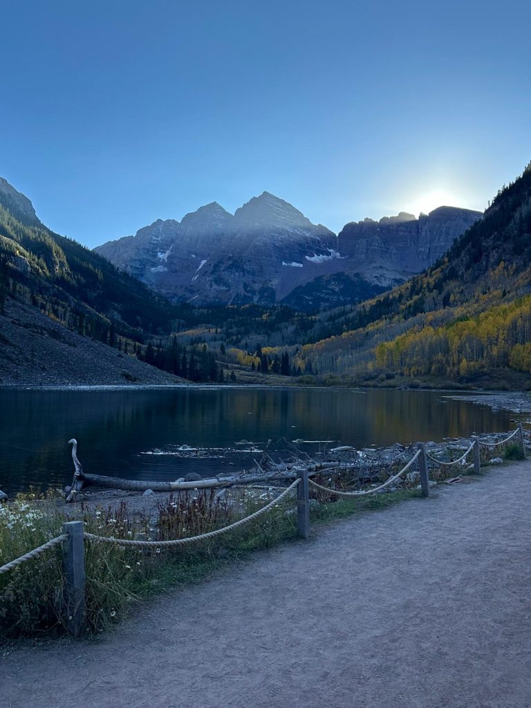 This photo showcases a serene mountain landscape in Maroon Bells, Aspen, during what appears to be early morning or late afternoon, as the sun is just beginning to rise or set behind the towering mountain peaks. The mountains are snow-capped and cast long shadows, adding depth to the scene. In the foreground, there is a calm, reflective lake bordered by a path lined with wooden posts and ropes, giving the area a natural, rustic feel.