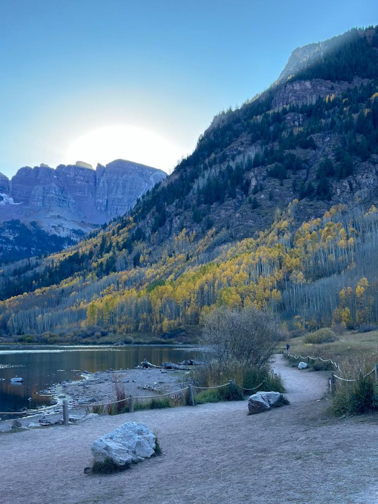 This photo captures a stunning mountain landscape with a lake in the foreground and tall, rugged peaks in the background. The sun is just starting to rise or set behind the mountains, creating a glowing halo of light around the peaks, giving the scene a dramatic and peaceful feel. The light from the sun softly illuminates the golden-yellow aspens on the hillside, signaling the vibrant colors of autumn. The lake reflects the surrounding scenery, and a natural path winds along its edge, bordered by ropes and dotted with large rocks and bushes. The path leads off into the distance, where a lone figure can be seen walking, adding a sense of scale to the majestic landscape. The mountainsides are a mix of evergreens and the golden aspens, with the contrast between the rocky terrain and the lush foliage enhancing the depth of the image. The overall atmosphere is tranquil, with the soft light and peaceful setting making it a serene and inviting scene.