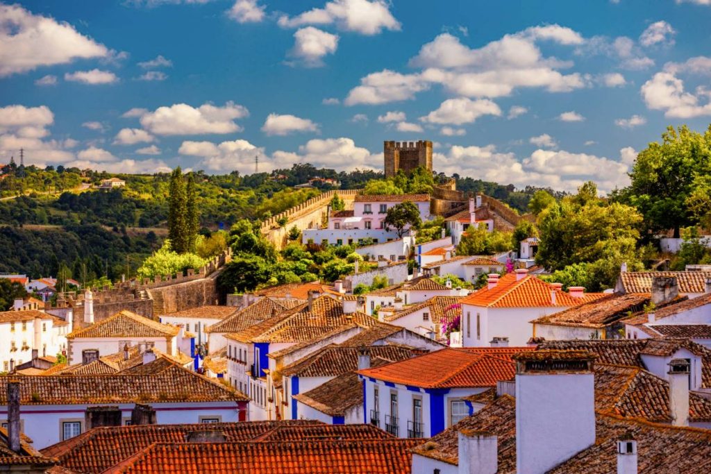 A panoramic view of Óbidos, Portugal, showcasing the traditional white buildings with terracotta roofs nestled closely together. The town is surrounded by lush greenery and trees, with a prominent medieval castle tower rising above on a hill under a partly cloudy sky. One of the Highlights of Portugal and a must see in any Curated Travel Itinerary.