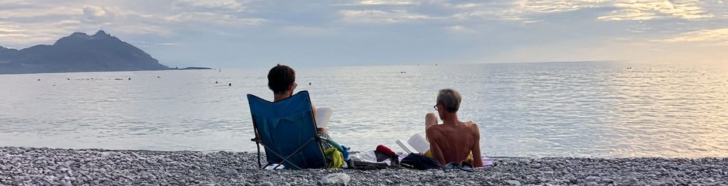 Two people sitting on a Camogli´s beach by the calm Ligurian sea, one in a blue folding chair and the other on a towel, both reading books. The scene is tranquil with a mountainous silhouette in the background and a soft, cloudy sky above.