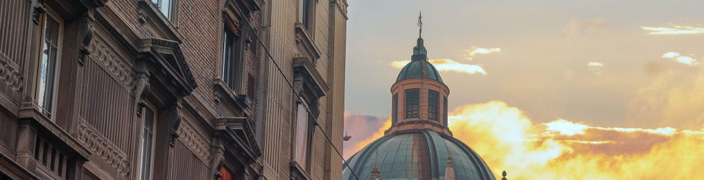 The image shows a picturesque urban scene at sunset, featuring the historical Bologna cityscape. A large domed building with a greenish-blue roof dominates the center, partially lit by the warm hues of the setting sun. The surrounding buildings are adorned with classical architecture, detailed facades, and shuttered windows. The foreground features ornate wrought-iron balcony railings and the street appears narrow, creating a cozy, old-town atmosphere. Snow dusts the rooftops, hinting at a winter setting.