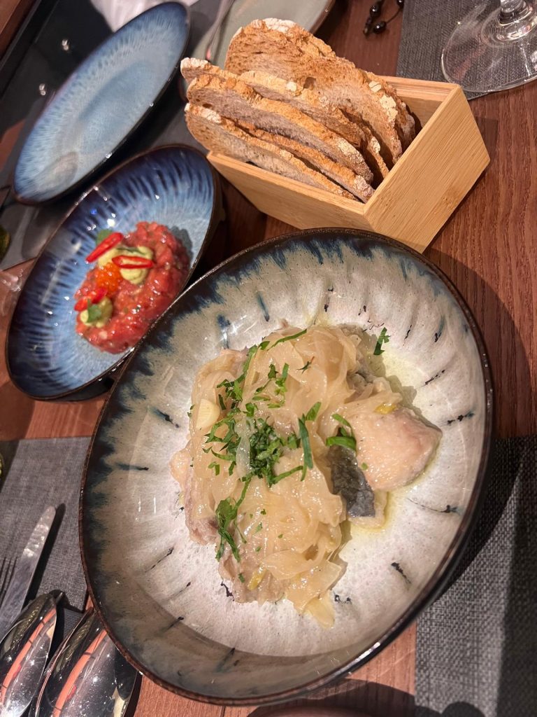 Photo of two starters served at Bistro restaurant in Matosinhos. On one plate is escabeche de carapau, a traditional Portuguese dish featuring marinated mackerel with vibrant colors and a tangy, aromatic presentation. The other plate showcases a delicately arranged steak tartare, topped with a garnish and accompanied by crisp elements. The dishes are beautifully plated, reflecting the restaurant's blend of tradition and contemporary culinary artistry.