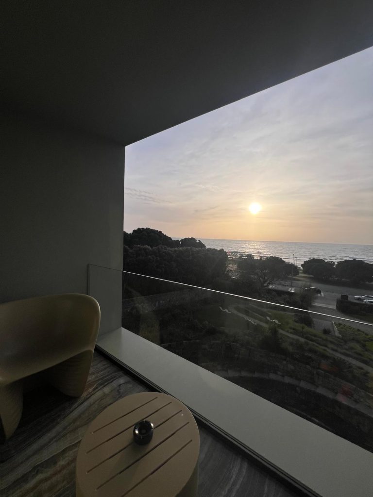 Photo of a stunning sunset viewed through a window at Vila Foz Hotel and Spa. The sky is painted in hues of orange with the sun dipping below the horizon. The silhouette of the window frame adds depth to the image, while the soft light from the sunset bathes the room in a warm, tranquil glow, enhancing the serene and luxurious ambiance.