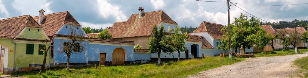 The image shows a picturesque rural village in Romania with traditional houses painted in pastel colors like yellow and blue, featuring steep red-tiled roofs. A dirt road runs through the scene, bordered by greenery, small trees, and open grassy areas. The sky above is bright with scattered clouds, adding to the tranquil, countryside atmosphere
