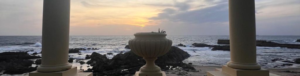A scenic view of the ocean at sunset, framed by two classical columns and a decorative urn in the foreground, with rocky shores and gentle waves under a soft, cloudy sky.