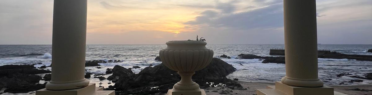 A scenic view of the ocean at sunset, framed by two classical columns and a decorative urn in the foreground, with rocky shores and gentle waves under a soft, cloudy sky.