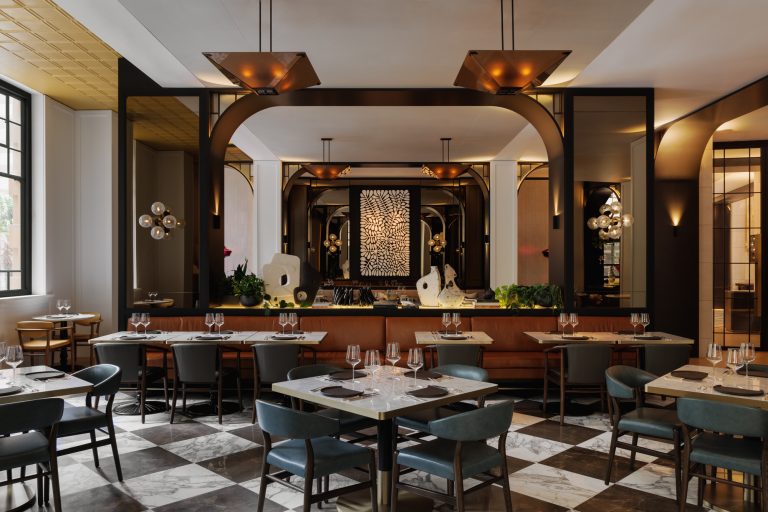 A refined dining room in Capella Sydney with a luxurious and modern design. The space features marble-tiled floors in a black-and-white checkerboard pattern, tables set with glassware and elegant table settings, and comfortable chairs upholstered in deep blue. The walls are accented with arched mirrors, creating a sense of depth and symmetry. Warm, ambient lighting from pendant fixtures and sconces enhances the sophisticated atmosphere. A centerpiece display includes abstract sculptures and lush greenery, with an intricate monochrome artwork framed prominently in the background.