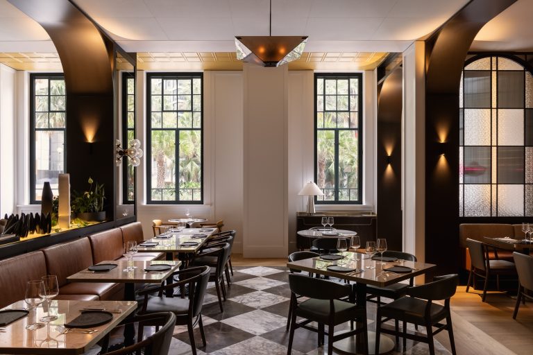 A refined dining room in Capella Sydney with a luxurious and modern design. The space features marble-tiled floors in a black-and-white checkerboard pattern, tables set with glassware and elegant table settings, and comfortable chairs upholstered in deep blue. The walls are accented with arched mirrors, creating a sense of depth and symmetry. Warm, ambient lighting from pendant fixtures and sconces enhances the sophisticated atmosphere. A centerpiece display includes abstract sculptures and lush greenery, with an intricate monochrome artwork framed prominently in the background.