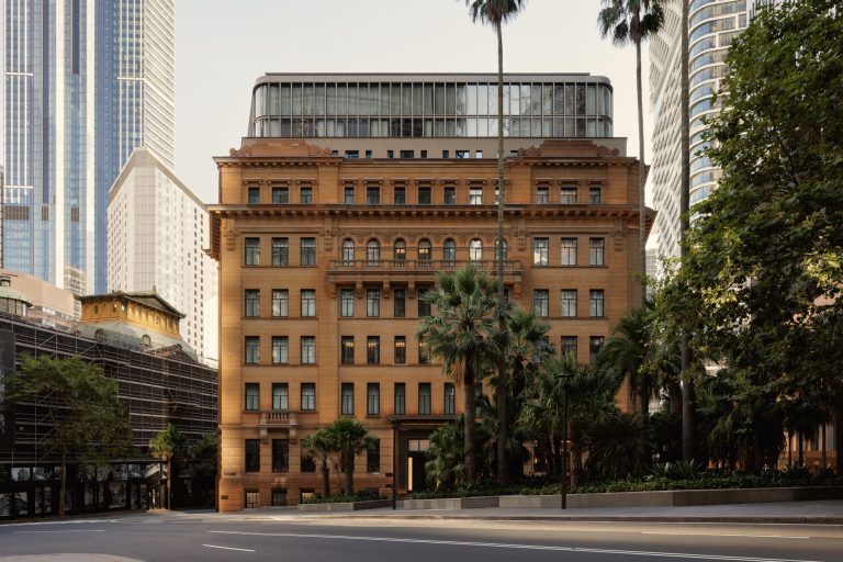 An elegant exterior view of Capella Sydney, showcasing its grand architecture that blends heritage charm with contemporary design. The building features ornate stonework, tall arched windows, and intricate detailing that reflects its historic origins. Lush greenery frames the scene, enhancing the luxurious ambiance. The soft lighting adds warmth, highlighting the sophistication and exclusivity of the landmark property.