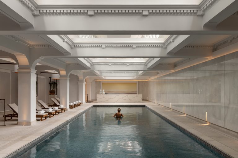 A young woman relaxes at an indoor pool within the Capella Sydney spa. The serene space features elegant architecture with soft lighting, celing windows, and sleek stone finishes. The tranquil water reflects the refined surroundings, while the woman sits on the pool's edge, wearing a white robe and gazing peacefully at the calm surface. A luxurious, calming atmosphere is evident, highlighting relaxation and sophistication.