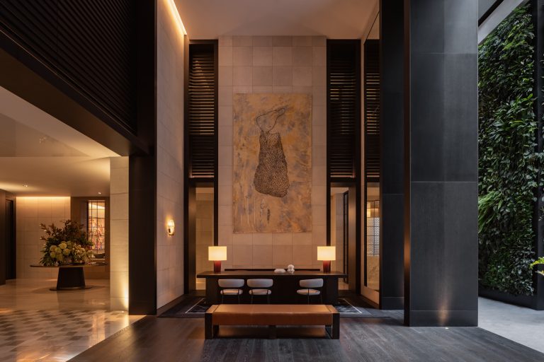 The reception area of Capella Sydney exudes luxury and sophistication. The space features high ceilings, warm ambient lighting, and a blend of modern and heritage-inspired design elements. A grand reception desk, crafted from polished stone, sits in front of a statement backdrop adorned with intricate patterns or artwork. Plush seating areas with elegant furnishings and rich textures are arranged nearby, inviting comfort and relaxation. The neutral color palette, accented by metallic and wood tones, enhances the refined atmosphere.