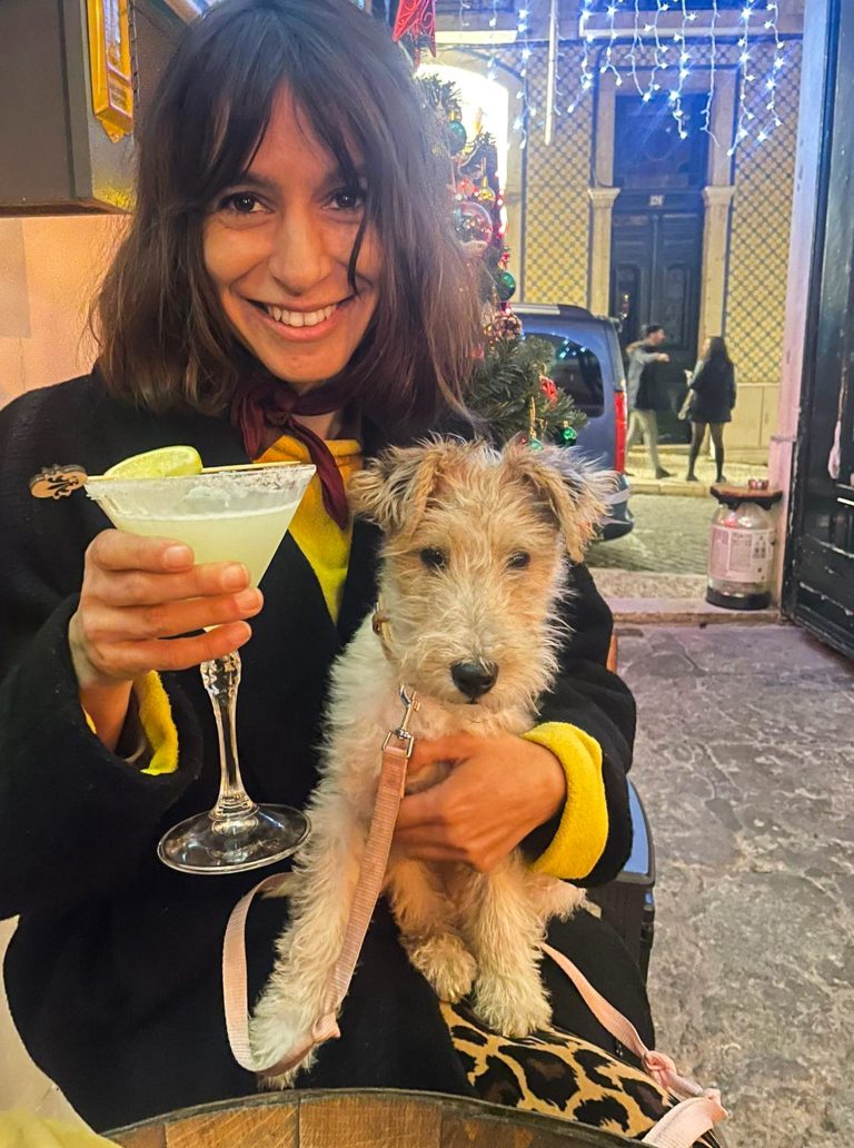 The image shows Cheila smiling warmly while holding Julião with light, curly fur. She is also holding a glass with a cocktail garnished with a slice of lime. The setting appears festive, with Christmas decorations, fairy lights, and a tree in the background. The scene takes place outdoors, possibly on a street with tiled walls and some people in the distance. The atmosphere feels cheerful and cozy.