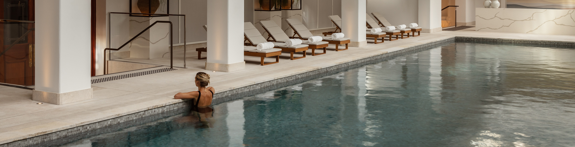 A young woman relaxes at an indoor pool within the Capella Sydney spa. The serene space features elegant architecture with soft lighting, and sleek stone finishes. The tranquil water reflects the refined surroundings, while the woman sits on the pool's edge. A luxurious, calming atmosphere is evident, highlighting relaxation and sophistication.