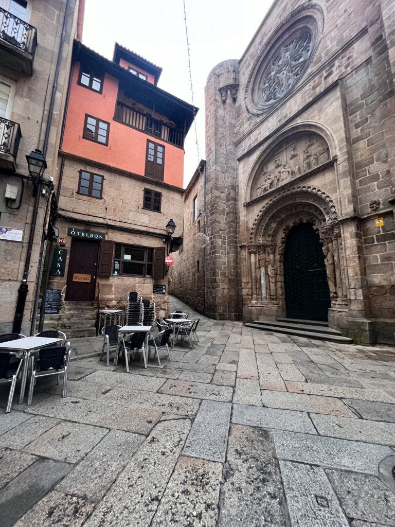 A street in Ourense, Spain, leads towards the Ourense Cathedral. The street is paved with light-colored stones and lined with old buildings. On the left, a restaurant named "O TRÉBON" has outdoor seating. In the distance, the large stone facade of the cathedral is visible, with its intricate carvings and a large rose window. The sky is overcast, and the light is soft.