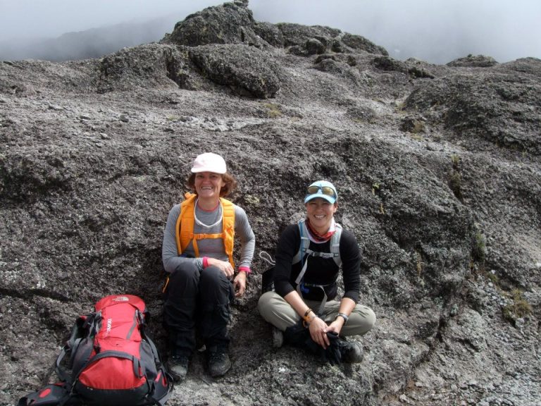 Cathy and Robin pause for a well-earned rest on a dark rock during their climb to Kilimanjaro, smiling with joy and pride at their achievement.