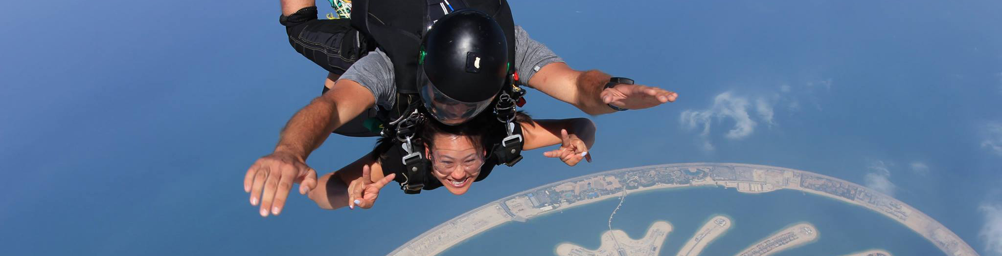 Cathy is free-falling from an airplane with a stunning aerial view of the Palm Jumeirah in Dubai beneath them. The instructor, wearing a black helmet and harness, is securely attached to Cathy, who is smiling and making a peace sign with her fingers. The deep blue sky and ocean provide a breathtaking backdrop, while the artificial island's unique palm shape is visible in the background.