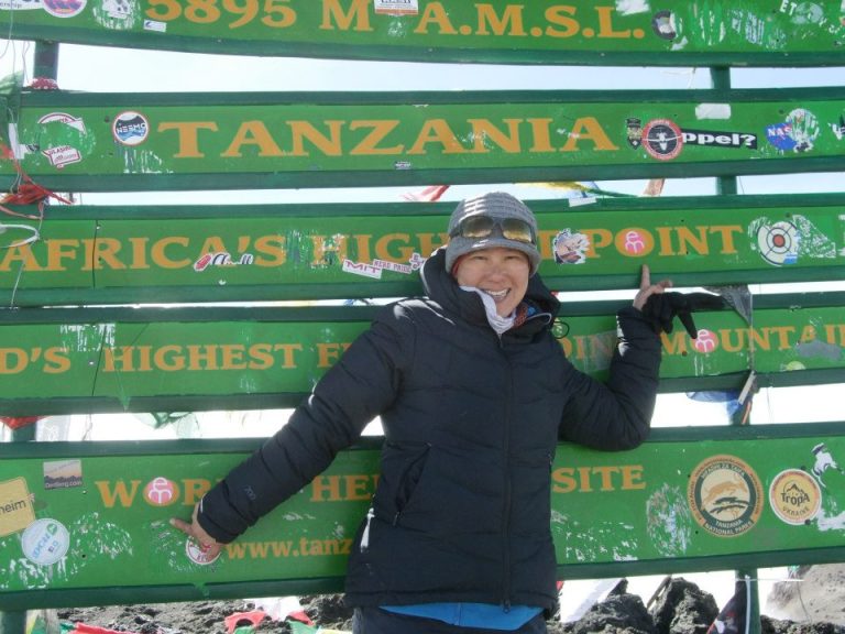 Cathy stands in front of a billboard on her journey to the highest peak of Mount Kilimanjaro.