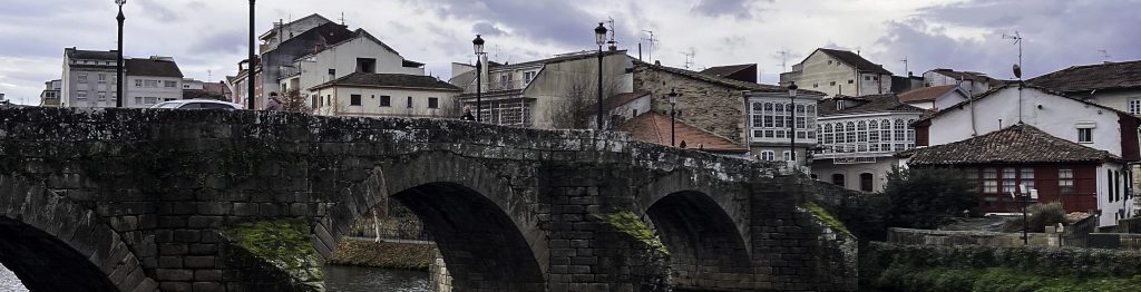 Scenic view of the medieval stone bridge over the Cabe River in Monforte de Lemos, Spain. The historic bridge features multiple arches covered in moss, reflecting in the calm waters. In the foreground, a red bench sits on a grassy area with fallen leaves, while leafless trees frame the scene. The background showcases traditional Galician buildings with white and red facades under a cloudy sky.