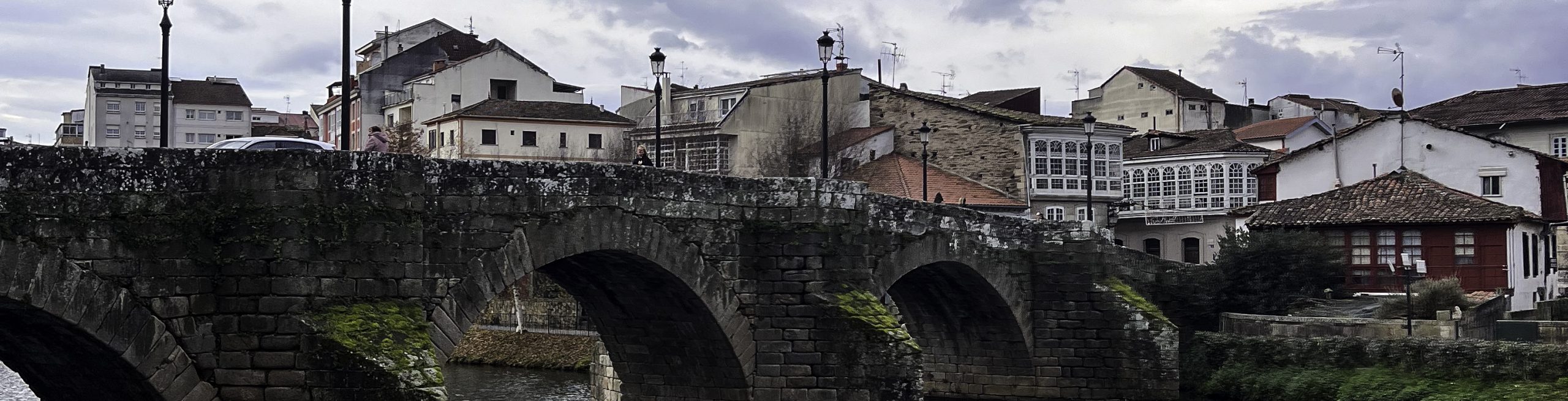 Scenic view of the medieval stone bridge over the Cabe River in Monforte de Lemos, Spain. The historic bridge features multiple arches covered in moss, reflecting in the calm waters. In the foreground, a red bench sits on a grassy area with fallen leaves, while leafless trees frame the scene. The background showcases traditional Galician buildings with white and red facades under a cloudy sky.