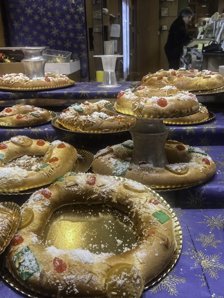 A display case filled with Roscón de Reyes, a traditional Spanish sweet bread eaten during the Christmas season. The cakes are round with a hole in the center and decorated with candied fruit and powdered sugar. The cakes are displayed on golden platters on a blue tablecloth with gold snowflake designs