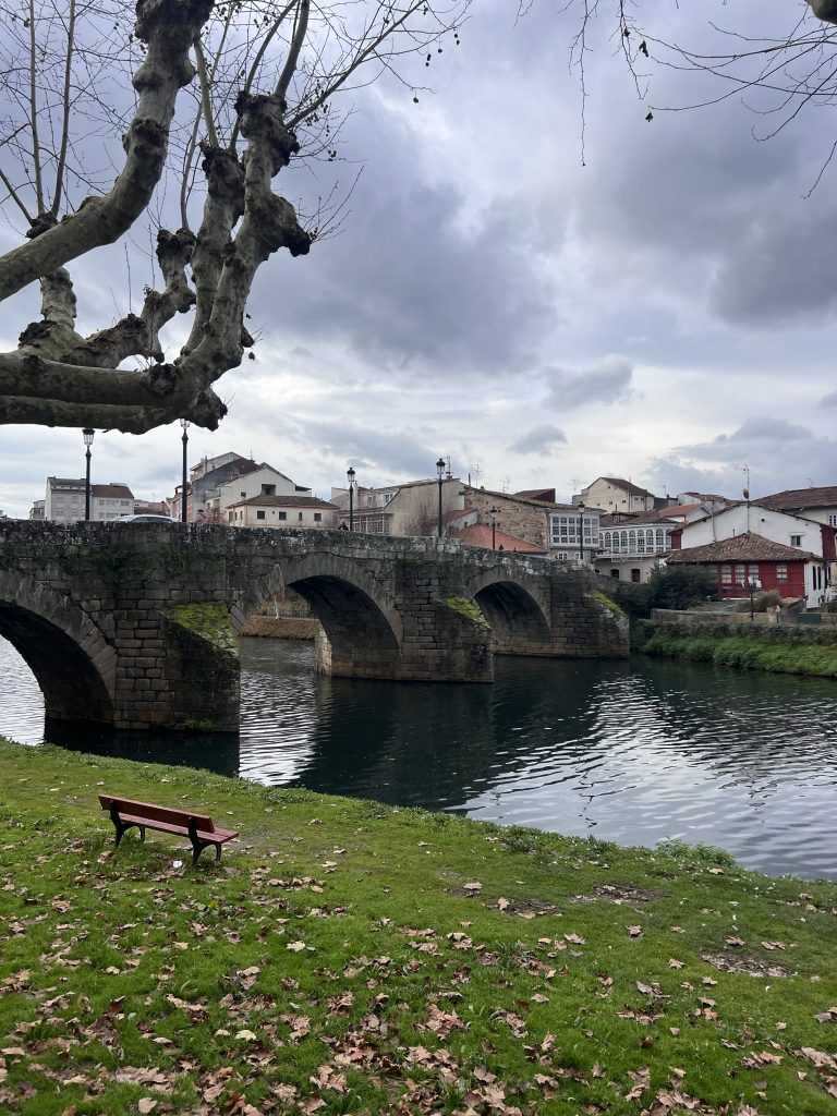 cenic view of Monforte de Lemos, Spain, featuring a historic stone bridge with multiple arches crossing the calm waters of the Cabe River. A red bench sits on a grassy riverbank covered with fallen leaves, while leafless trees frame the cloudy sky. Traditional Galician buildings with white and red facades line the background.