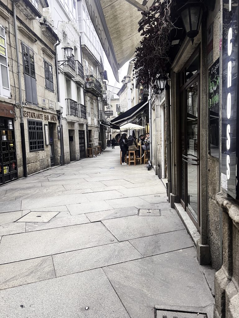 A street in Lugo, Spain, with old stone buildings on either side. The street is paved with large, light-colored stones and is narrow, with barely enough room for a car to pass. There are a few people walking on the street, and some tables and chairs are set up outside a restaurant. The buildings are tall and narrow, with balconies and windows overlooking the street. The sky is overcast and the light is dim