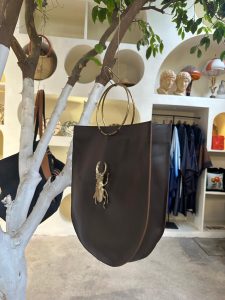 A dark brown leather tote bag with a distinctive gold-toned scarab beetle embellishment is the central focus of the image. The bag has a rounded bottom and is suspended by two large, circular gold metal handles from a thin gold chain or hook attached to a white-painted tree branch. The tree branch extends diagonally across the frame, with a few green leaves visible at the top.