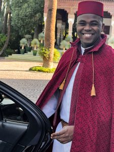 A smiling man stands next to a black car, wearing a traditional red fez hat and a patterned red cape with gold tassel ties. He has a light complexion and short, dark hair. He is wearing a white garment underneath the cape and a woven belt around his waist. The man is looking towards the right side of the frame with a cheerful expression, showing his white teeth. The background features a paved area, lush green landscaping with palm trees and other greenery, and a glimpse of an ornate building with archways in the distance.