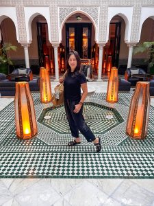 Cathy smiles at the camera while standing in a beautifully tiled courtyard. She is wearing a short-sleeved black shirt, black pants rolled up at the ankles, and black sandals. A light brown patterned tote bag hangs from her left shoulder.
