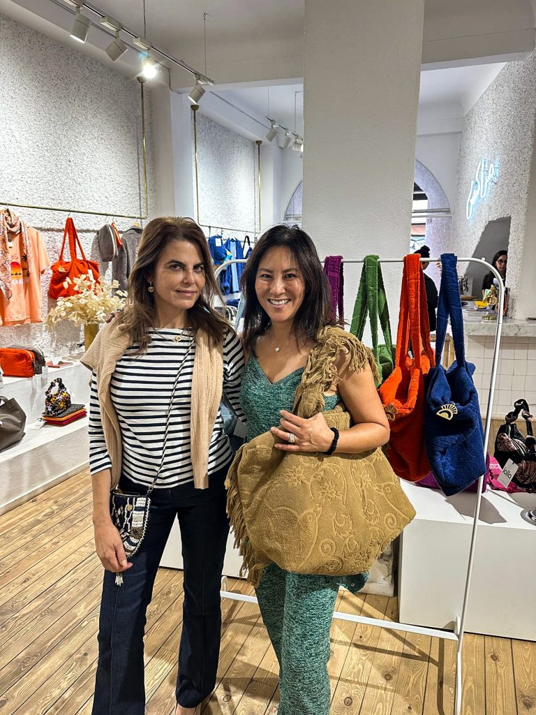 Cathy and Mayan pose for a photo inside a brightly lit retail store. Maryan has shoulder-length brown hair and is wearing a black and white striped long-sleeved shirt, dark jeans, a light beige sweater draped over her shoulders, and a small, dark, embellished purse with a shoulder strap. She is also wearing earrings and a necklace. Cathy has shoulder-length dark hair and is wearing a green patterned outfit and a large, light brown, patterned tote bag with fringe over her shoulder. She is also wearing rings and a necklace.