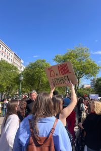 A person holds a cardboard sign reading '25 DE ABRIL SEMPRE!' above a crowd gathered outdoors in a sunny city square lined with trees and buildings.