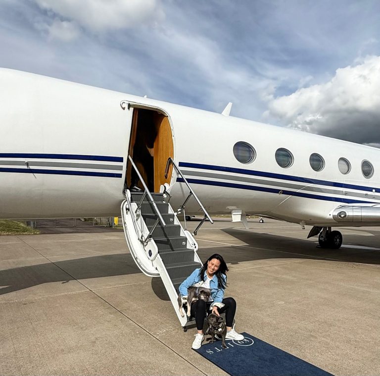 Cathy sitting on the private jet staircase with tbeluga and Luna, the two French bulldogs, on a K9 Jets-branded mat, with the jet and partly cloudy sky in the background.