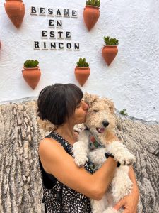 A vertical, medium close-up shot of Cheila, holding and kissing her dog, Juliao. Cheila has her hair in a bun and is wearing a black and white patterned top. She is holding the fluffy, light-colored dog with both arms and is leaning in to kiss its cheek. Juliao is looking at the camera with his mouth slightly open, appearing to smile. In the background, a white wall is decorated with five terracotta cone-shaped planters holding small succulent plants. The Spanish text "BESAME EN ESTE RINCON" (Kiss me in this corner) is written on the wall in black tiles. A rocky, textured wall is visible in the foreground below them
