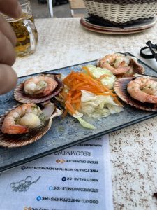 A top-down shot of a plate of zamburiñas (queen scallops) with shrimp, served on a restaurant table. The scallops are arranged on their shells with a large shrimp on top of each, garnished with herbs. In the center of the plate is a small salad of shredded carrots and lettuce. A menu with Spanish text is partially visible below the plate. A glass of beer and a hand are in the upper left corner of the image, and a woven bread basket is in the upper right