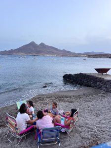 A group of four people and two children are seated in beach chairs on a dark, sandy beach at dusk, gathered around a low table. The people are facing the water, which is dotted with anchored boats. In the background, a large, dark mountain rises from the water, and the sky is a light blue with a few clouds. A small, rocky jetty is visible on the right side of the image. The light is fading, giving the scene a soft, gentle feel.