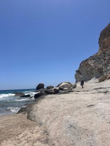 A vertical wide shot of Cala de Enmedio on a sunny day. The foreground shows a rocky, light-colored beach with waves lapping at the shore. The beach is bordered on the right by a large cliff face. The background is dominated by a clear blue sky. A few people are visible on the beach