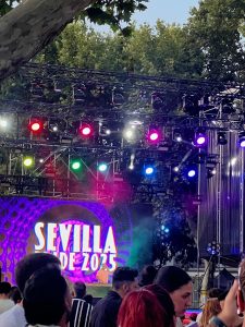A medium shot of an outdoor music festival or event at dusk. A large stage is set up with a truss structure and multiple bright, multi-colored stage lights. A large LED screen on the stage displays the words "SEVILLA DE 2025" in large, bold, purple letters. The heads and backs of a crowd are visible in the foreground, facing the stage. The event is taking place under a canopy of green trees.