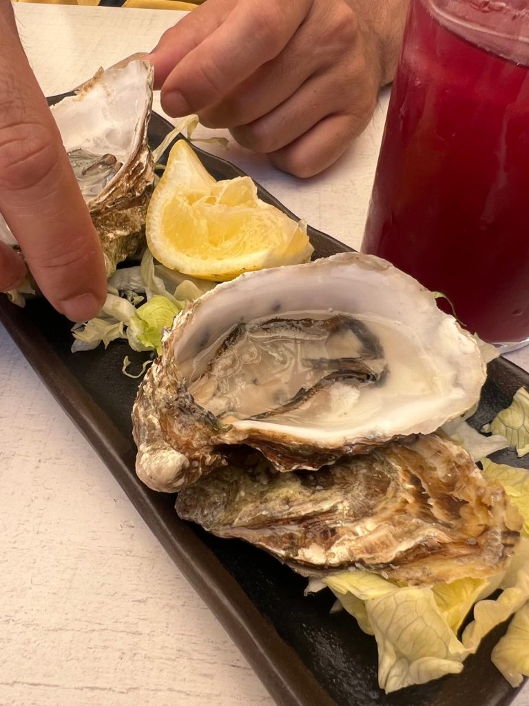 A close-up shot of a plate of fresh oysters. Three large oysters on the half-shell are arranged on a bed of shredded lettuce on a long, dark platter. A lemon wedge is placed in the middle of the plate. A person's hand is visible on the left, reaching towards one of the oysters. A glass containing tinto de verano with ice is partially visible in the top right corner. The table is a white, wooden surface.